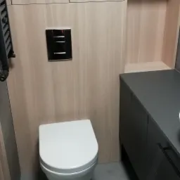 Nowoczesna toaleta podwieszana z chromowanym przyciskiem spłukiwania, obudowana jasnym drewnem i szarą szafką umywalkową z czarnymi uchwytami, na szarej podłodze.