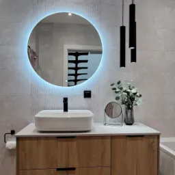 Nowoczesna łazienka z podświetlanym okrągłym lustrem, umywalką nablatową i szafką z drewnopodobnymi frontami, uzupełniona minimalistycznymi lampami wiszącymi i dekoracyjnym wazonem z zielenią.