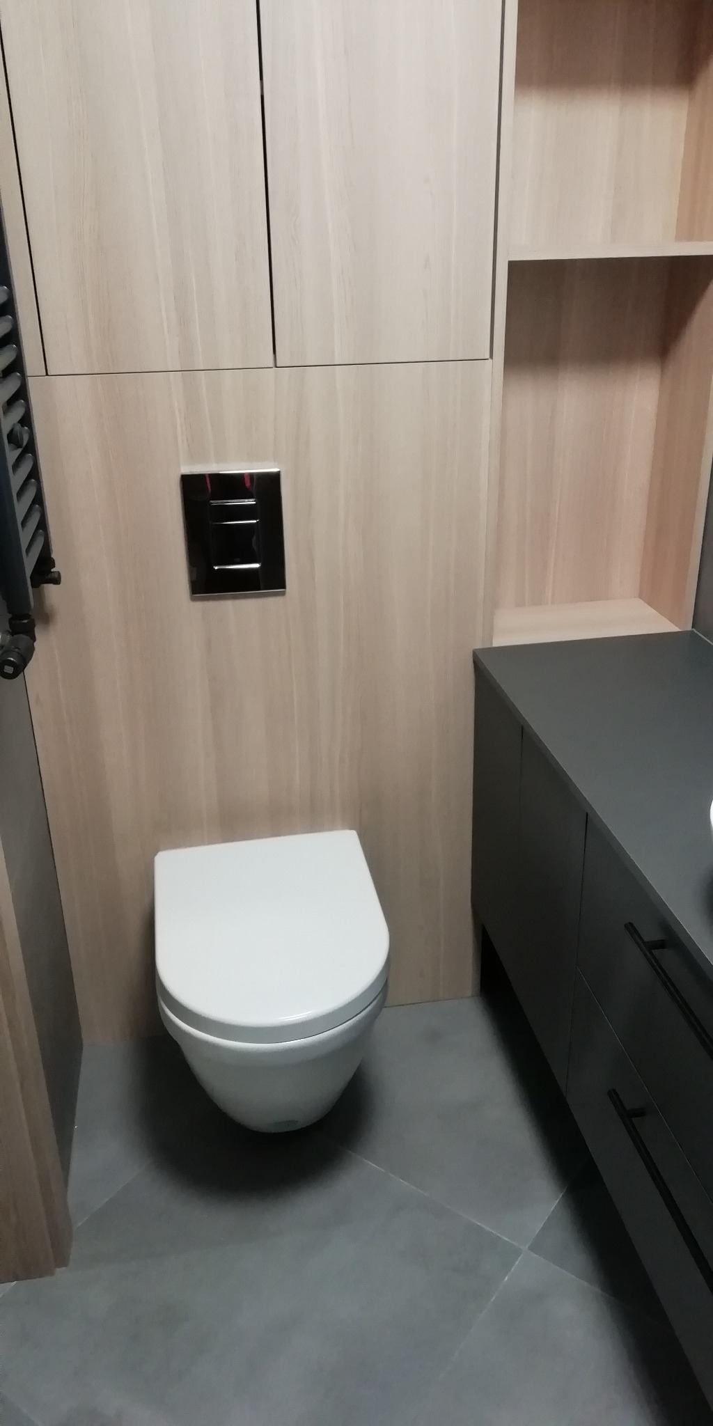 Nowoczesna toaleta podwieszana z chromowanym przyciskiem spłukiwania, obudowana jasnym drewnem i szarą szafką umywalkową z czarnymi uchwytami, na szarej podłodze.