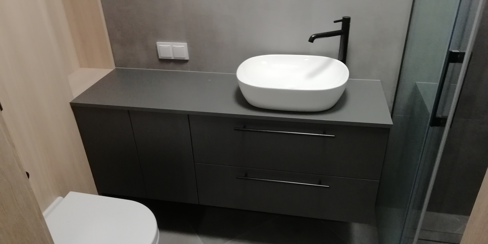 Szara szafka łazienkowa z dwiema szufladami i białą, owalną umywalką nablatową oraz czarną armaturą na tle szarej ściany i fragmentu kabiny prysznicowej.