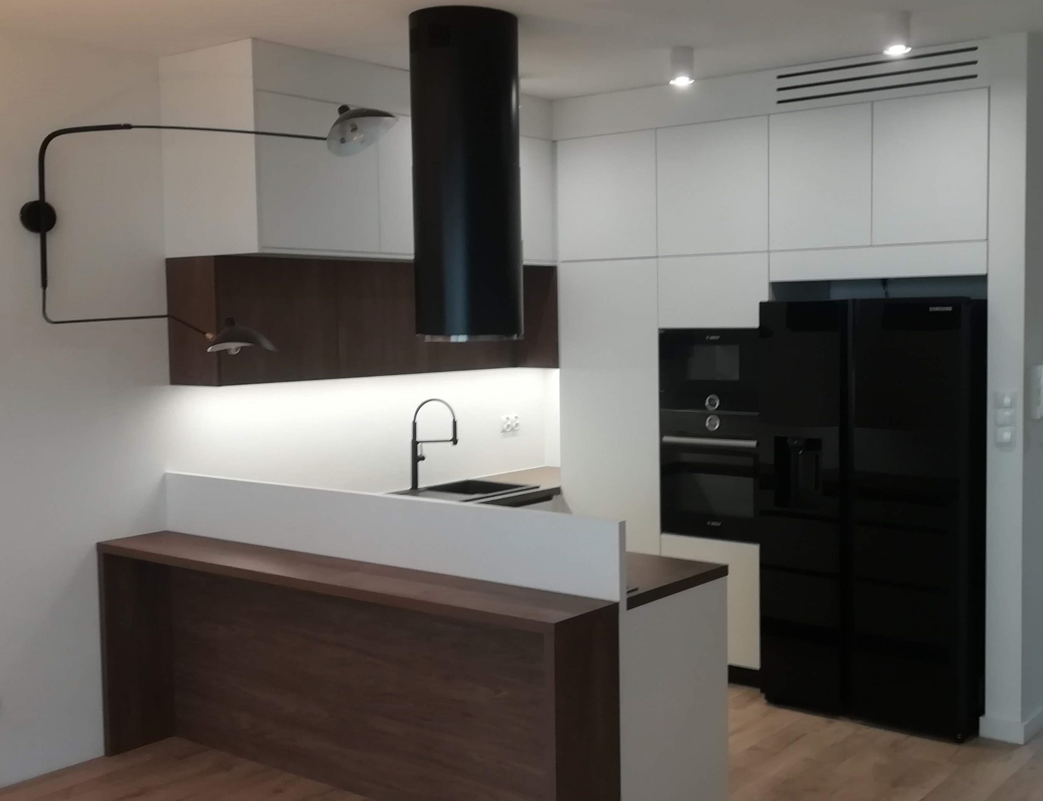 Nowoczesna kuchnia z białymi szafkami, brązowymi akcentami i czarnym wyposażeniem AGD, oświetlona designerską lampą ścienną.