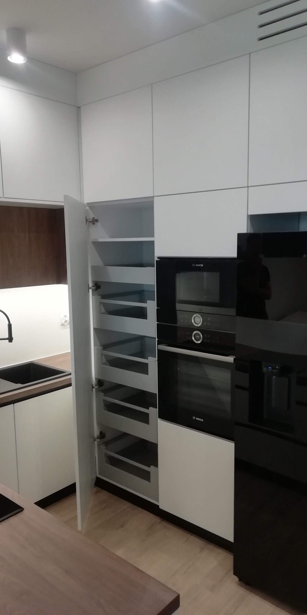 Nowoczesna, biała kuchnia z otwartą szafką typu cargo, ukazującą wysuwane półki; w tle zabudowa z piekarnikiem i mikrofalówką marki Bosch.