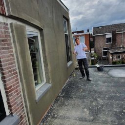 Kamil Sap Schildersbedrijf - Pracownik na dachu budynku po nałożeniu szarej warstwy tynku na ścianę, z widocznym oknem i fragmentem ceglanej elewacji, trzymający w ręku żółty wałek, z uniesionymi kciukami w górę.