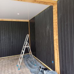 Kamil Sap Schildersbedrijf - Malowanie pionowych desek na czarno wewnątrz budynku, widoczna drabina, wiaderka z farbą i zabezpieczona podłoga.