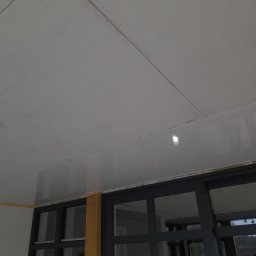 Kamil Sap Schildersbedrijf - Biały sufit podwieszany z widocznymi łączeniami płyt, częściowo pomalowany, z widoczną taśmą malarską przy oknie z ciemnymi ramami i fragmentem drabiny.