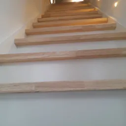 Jasne, drewniane schody z podświetleniem LED na ścianie, widziane z dołu, z fragmentem dywanu na dole kadru.