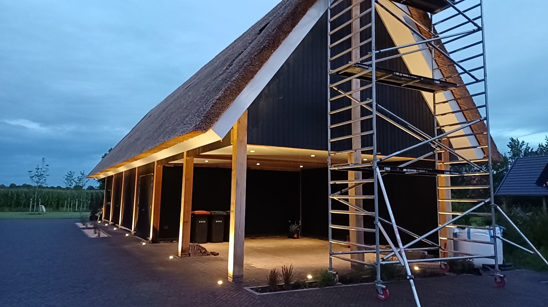 Budynek gospodarczy z ciemną elewacją i strzechą, oświetlony punktowymi lampami, z rusztowaniem przy ścianie bocznej.