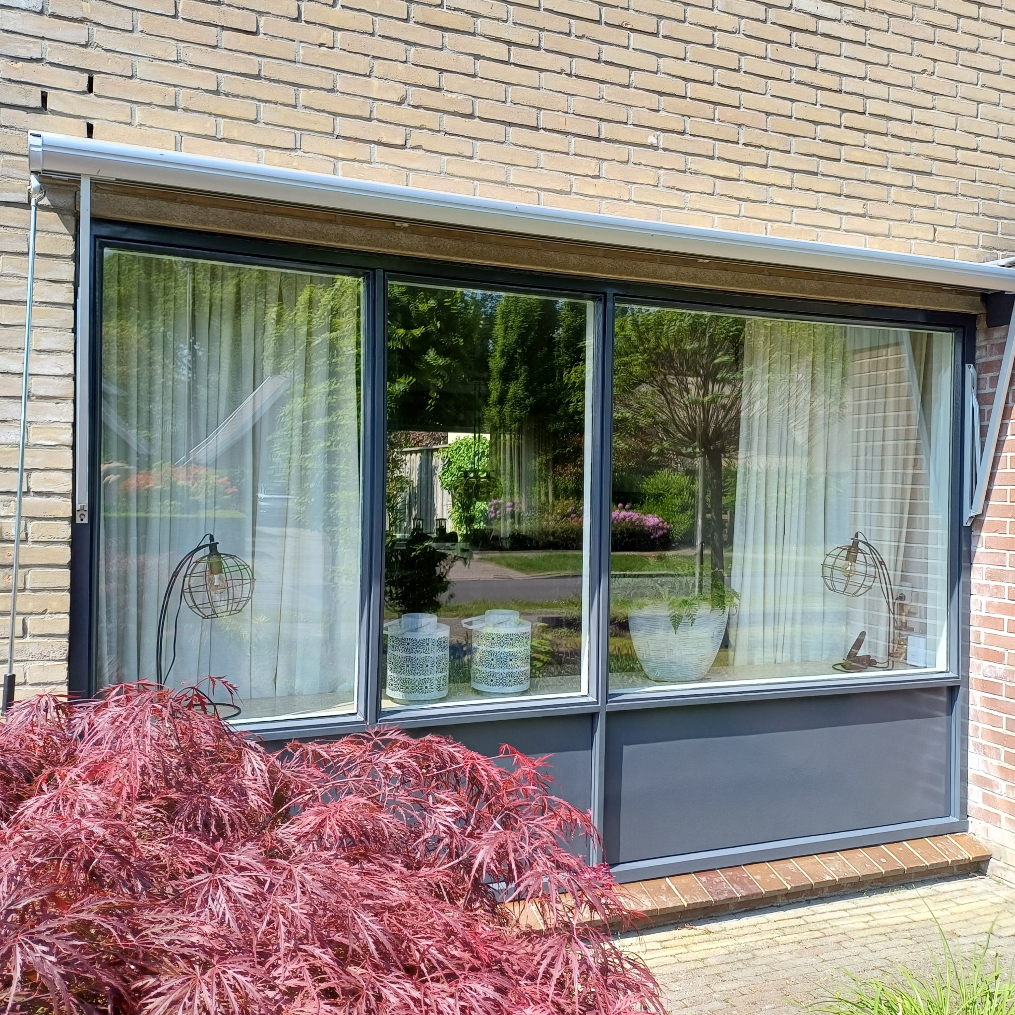 Trzyczęściowe okno z szarą ramą w ceglanym domu, zasłonięte firankami, z widokiem na ogród, dekorowane lampionami i lampą, z krzewem o bordowych liściach na pierwszym planie.