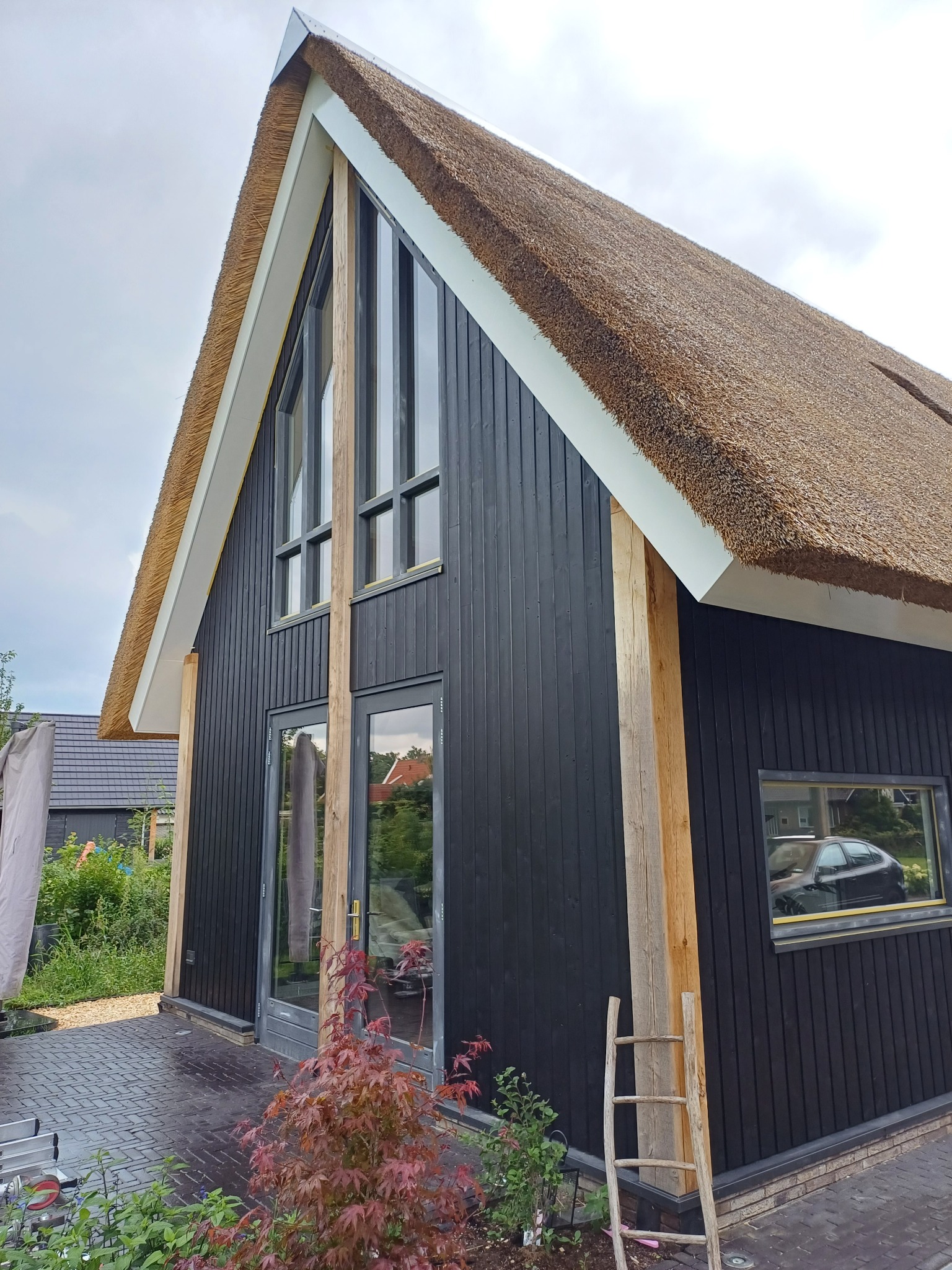 Nowoczesny dom z czarną drewnianą elewacją i strzechą, widoczne duże okna, drewniane belki konstrukcyjne, drabina oparta o ścianę i czerwony klon palmowy na pierwszym planie.