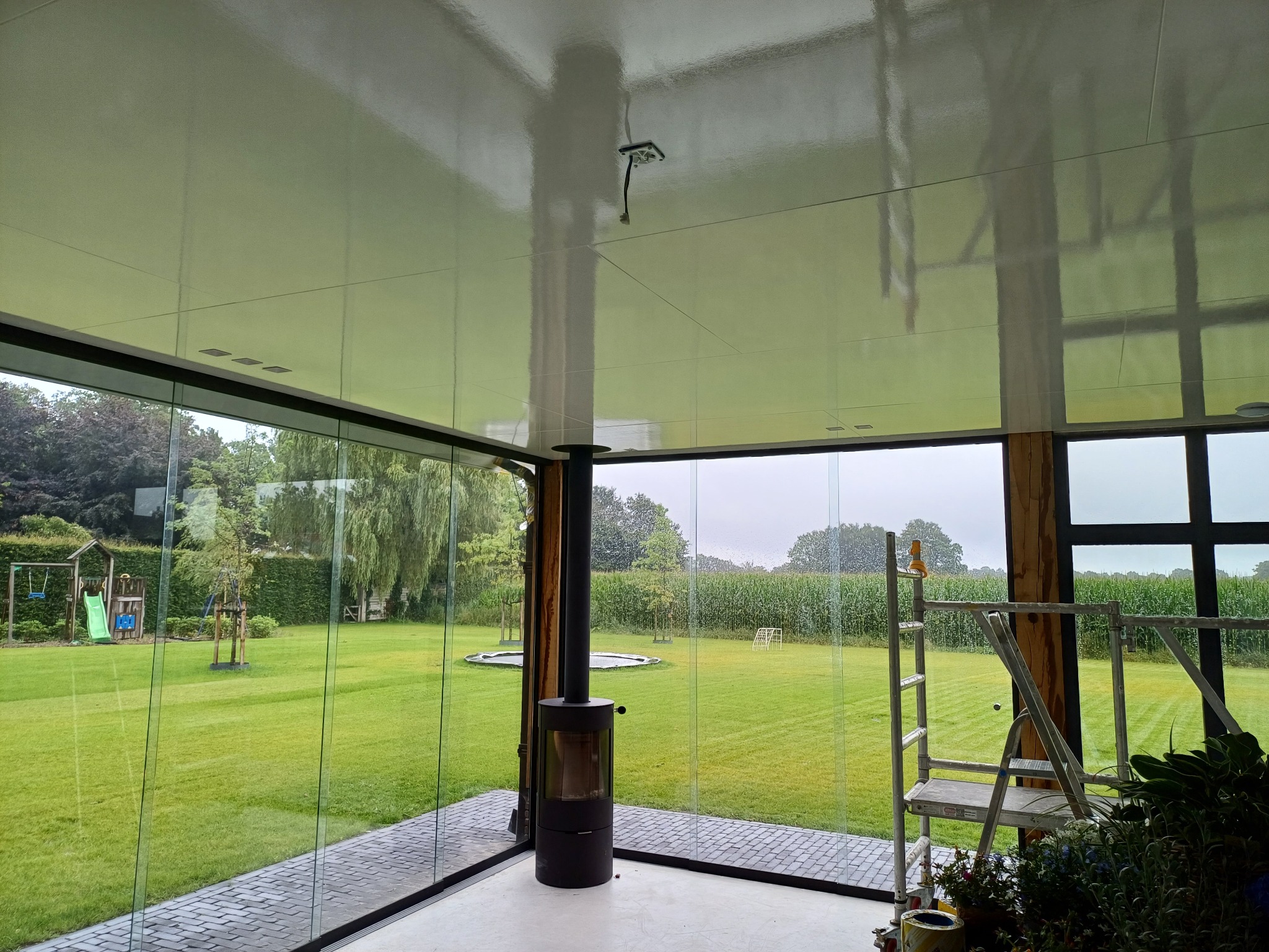Nowoczesna przeszklona weranda z widokiem na zadbany trawnik, czarny piec wolnostojący i aluminiowa drabina na kółkach wewnątrz.