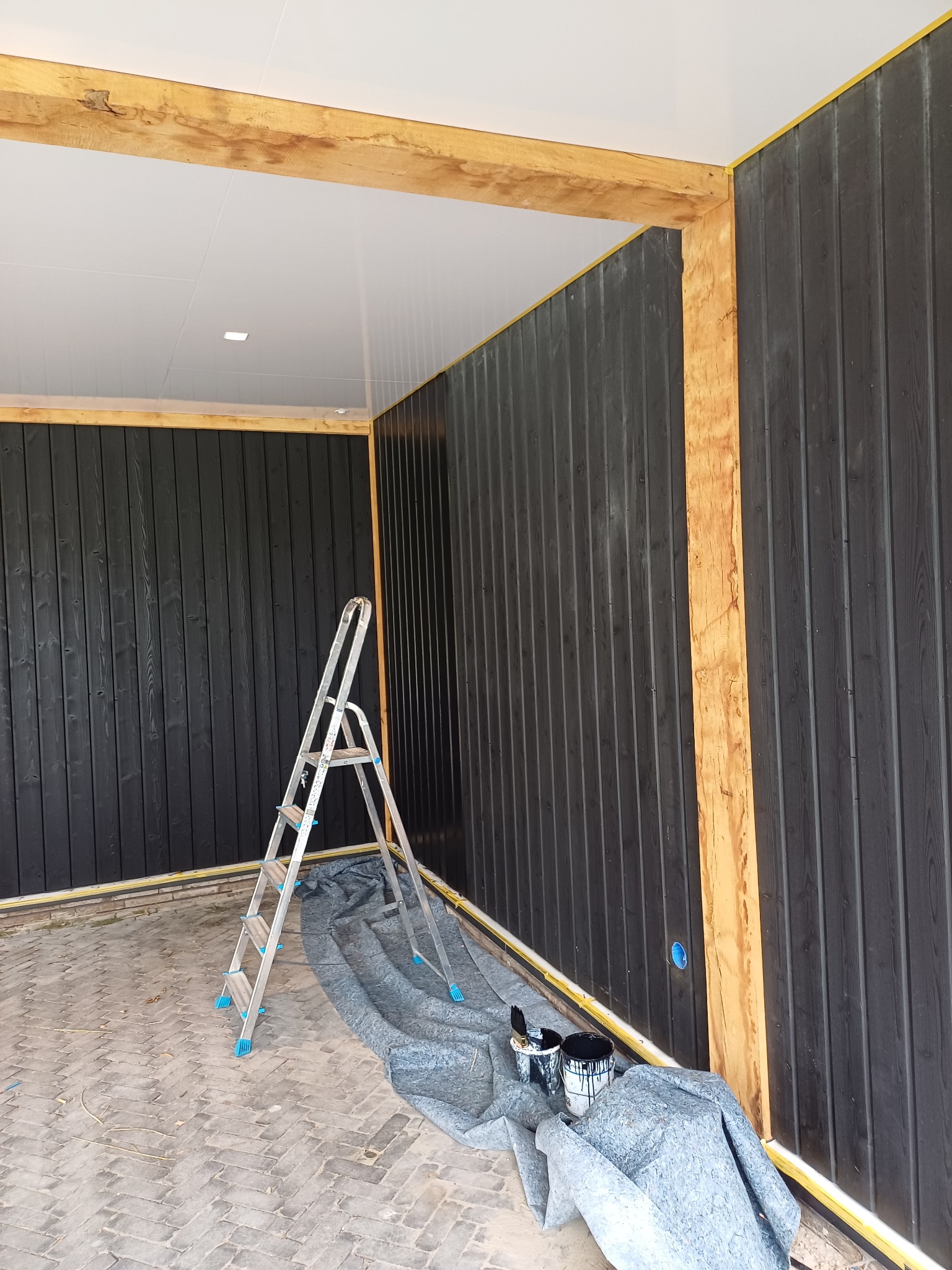 Malowanie pionowych desek na czarno wewnątrz budynku, widoczna drabina, wiaderka z farbą i zabezpieczona podłoga.