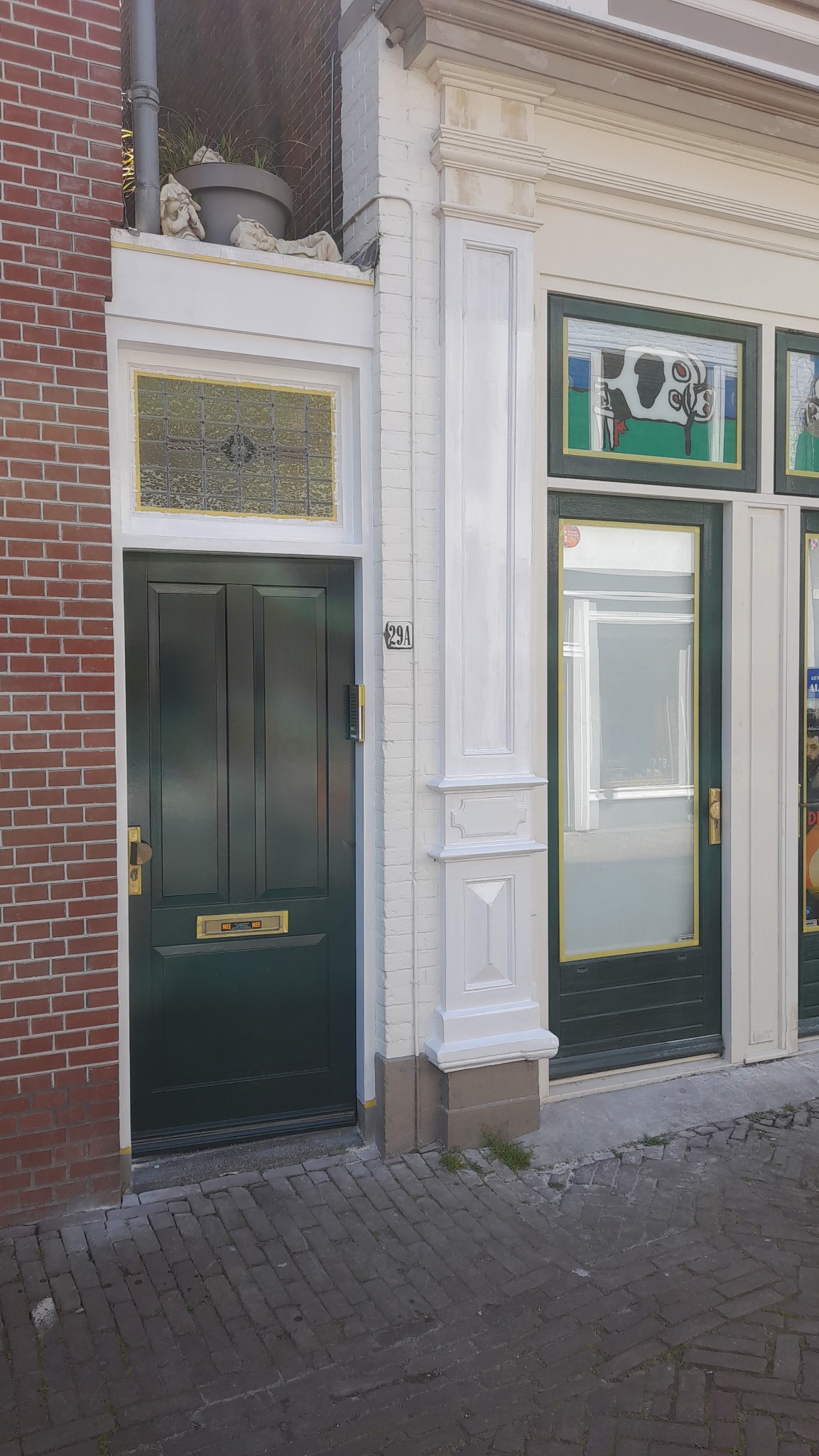 Fasada budynku z dwoma zielonymi drzwiami, numer 29A obok jednych z nich, ceglana ściana po lewej stronie i dekoracyjne elementy architektoniczne.