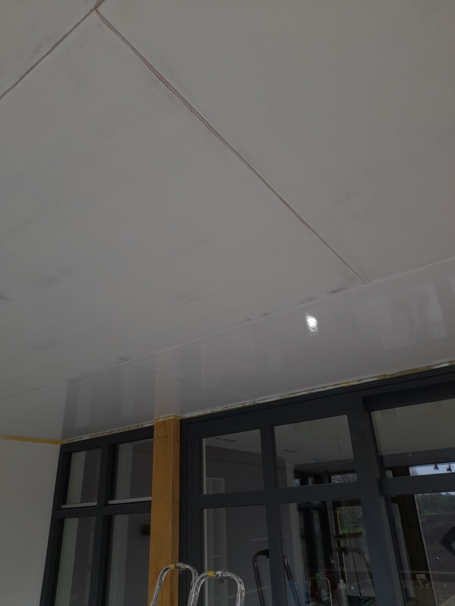 Biały sufit podwieszany z widocznymi łączeniami płyt, częściowo pomalowany, z widoczną taśmą malarską przy oknie z ciemnymi ramami i fragmentem drabiny.