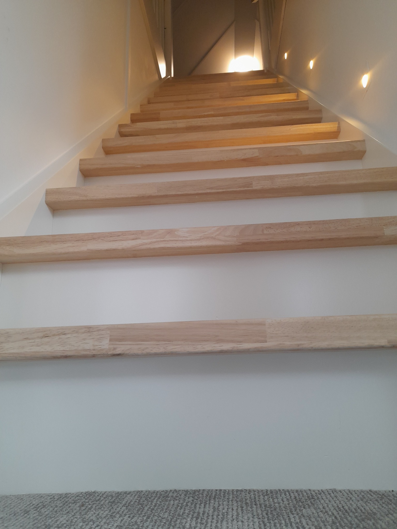 Jasne, drewniane schody z podświetleniem LED na ścianie, widziane z dołu, z fragmentem dywanu na dole kadru.