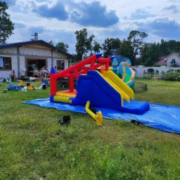 Duży, kolorowy dmuchany zamek ze zjeżdżalnią na trawniku, rozłożony na niebieskiej płachcie, w tle dom w trakcie budowy i bawiący się ludzie.