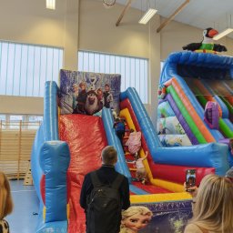 BAWEO BARBARA WÓJCIK - Kolorowy dmuchany zamek z motywem 'Kraina Lodu' w hali sportowej. Dzieci wspinają się po schodach, a dorośli robią zdjęcia. Na szczycie zjeżdżalni duży, dmuchany tukan.