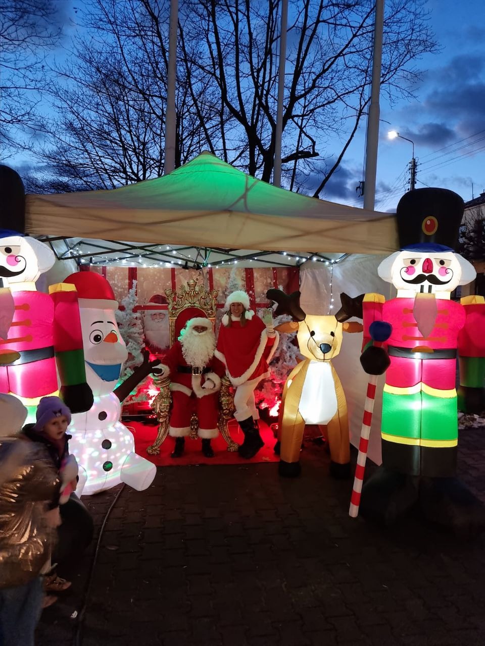 Świąteczna scena z dmuchanymi dekoracjami: Święty Mikołaj na tronie, bałwan Olaf, dziadek do orzechów i renifer, wszystko podświetlone na tle wieczornego nieba.