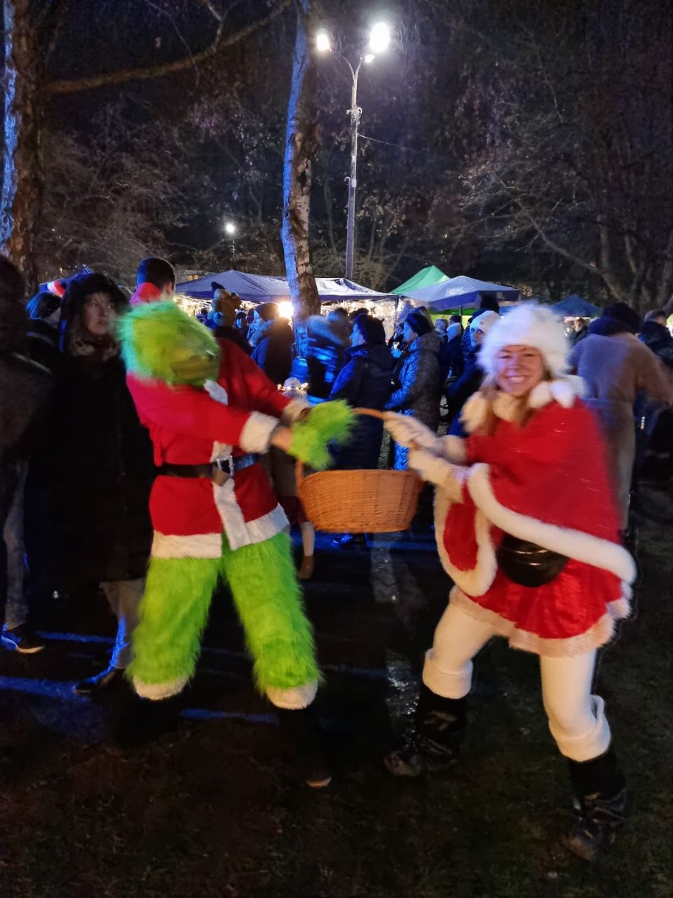 Animatorzy w kostiumach Grincha i Mikołajki, w plenerze, z koszykiem, podczas zabawy z publicznością na tle tłumu i oświetlenia.