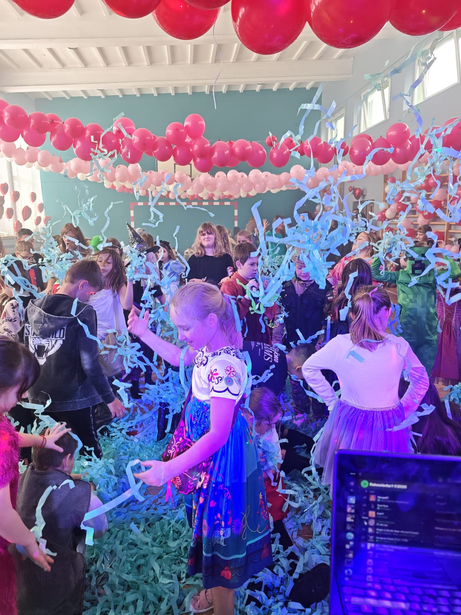 Dzieci bawią się w konfetti podczas imprezy z balonami. Dziewczynka w stroju księżniczki w centrum kadru. Laptop DJ-a w prawym dolnym rogu.