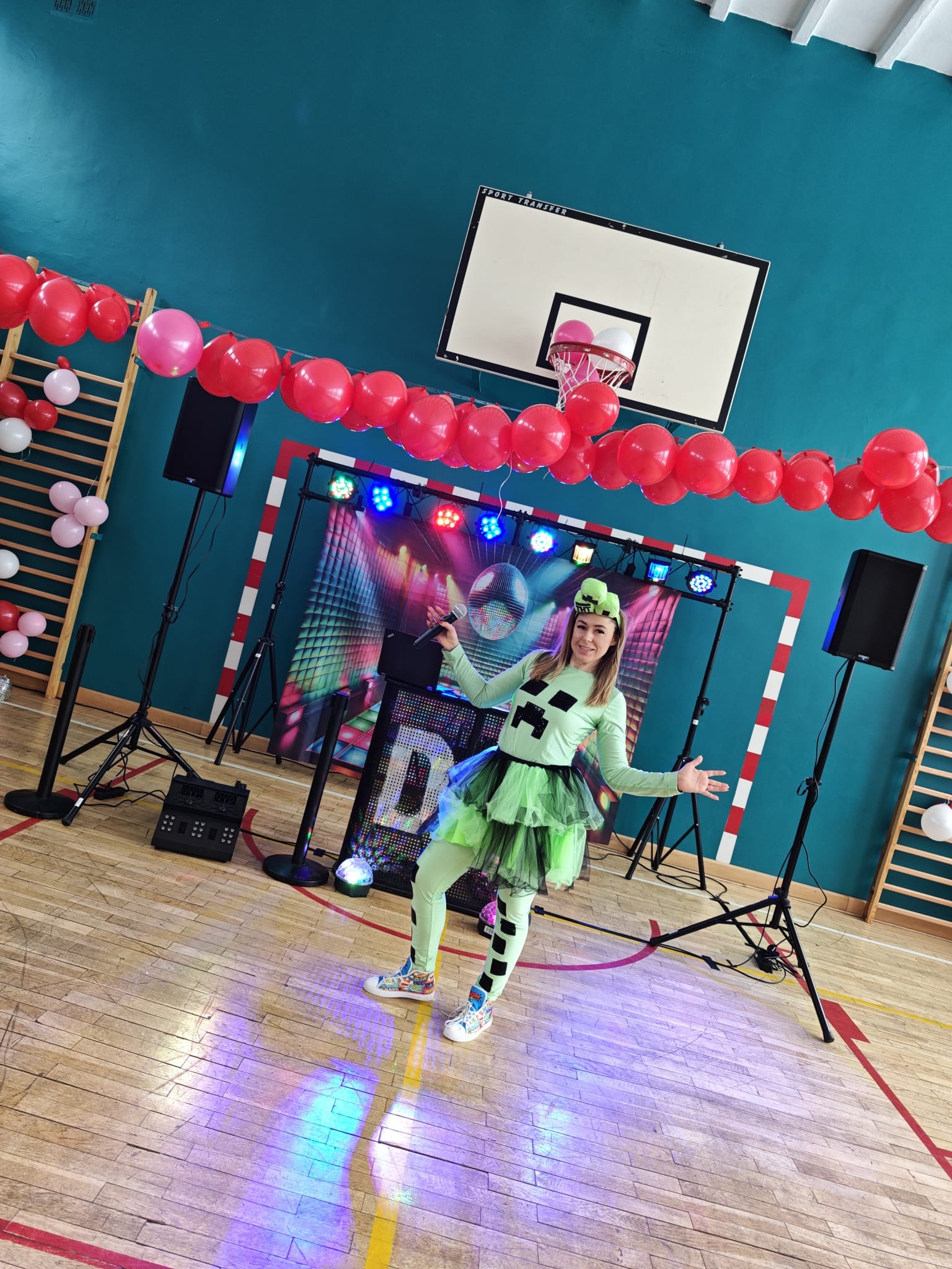 Animatorka w stroju Minecrafta z mikrofonem, na tle dekoracji z balonów i sprzętu nagłośnieniowego w sali gimnastycznej. Uśmiechnięta kobieta z rozłożonymi rękami.