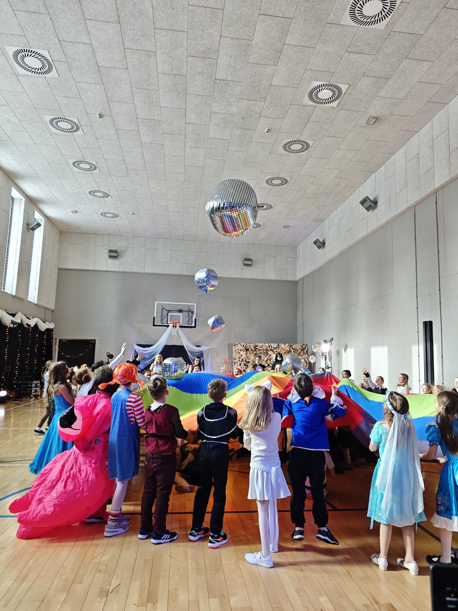 Dzieci w kostiumach bawią się kolorową chustą animacyjną w sali gimnastycznej, pod wiszącymi kulami dyskotekowymi. W tle scena z dekoracjami.