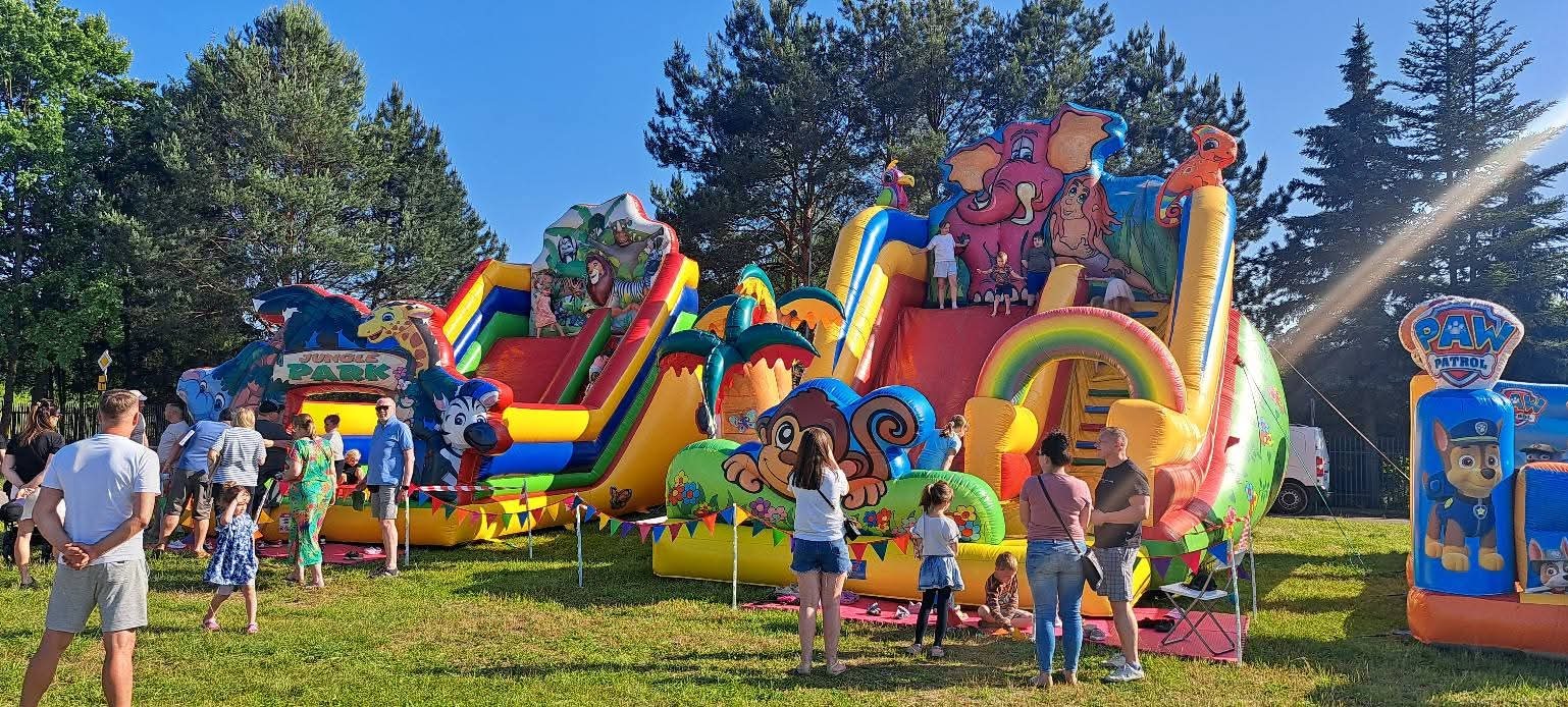 Kolorowe dmuchańce z motywem dżungli i Paw Patrol na zielonej trawie, otoczone grupą ludzi w słoneczny dzień. Widoczne drzewa w tle.