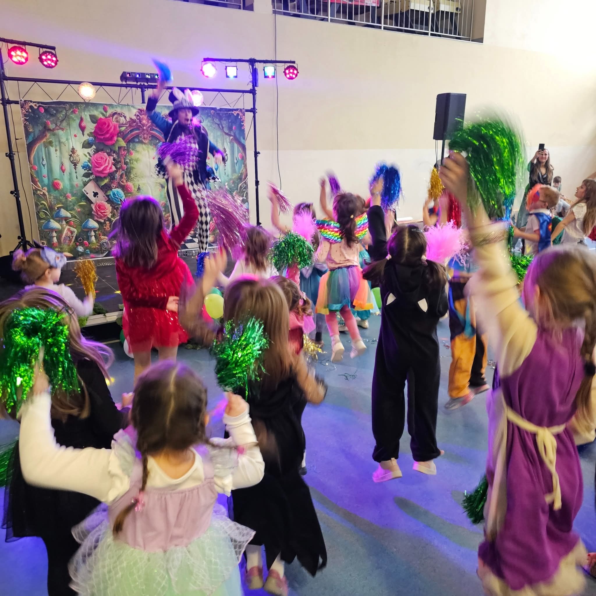 Animator w stroju Szalonego Kapelusznika prowadzi tańce z dziećmi z pomponami na tle scenografii z Alicji w Krainie Czarów, oświetlonej kolorowymi światłami.