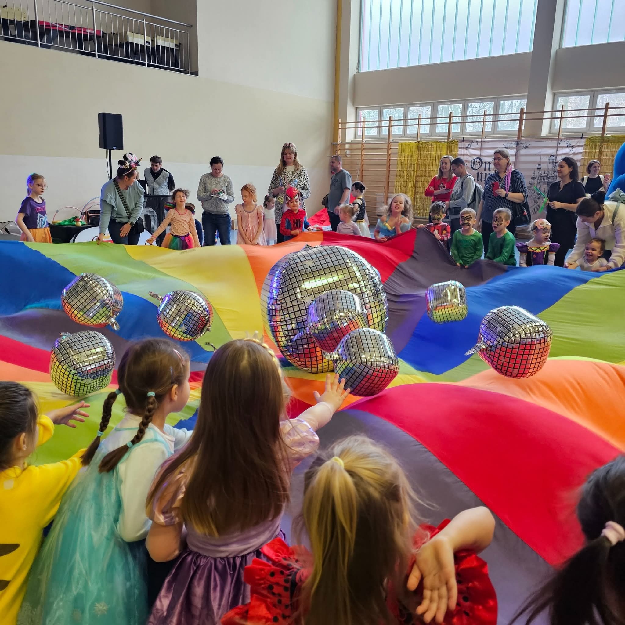 Dzieci bawią się kolorową chustą animacyjną z unoszącymi się srebrnymi kulami dyskotekowymi. W tle dorośli obserwują zabawę.