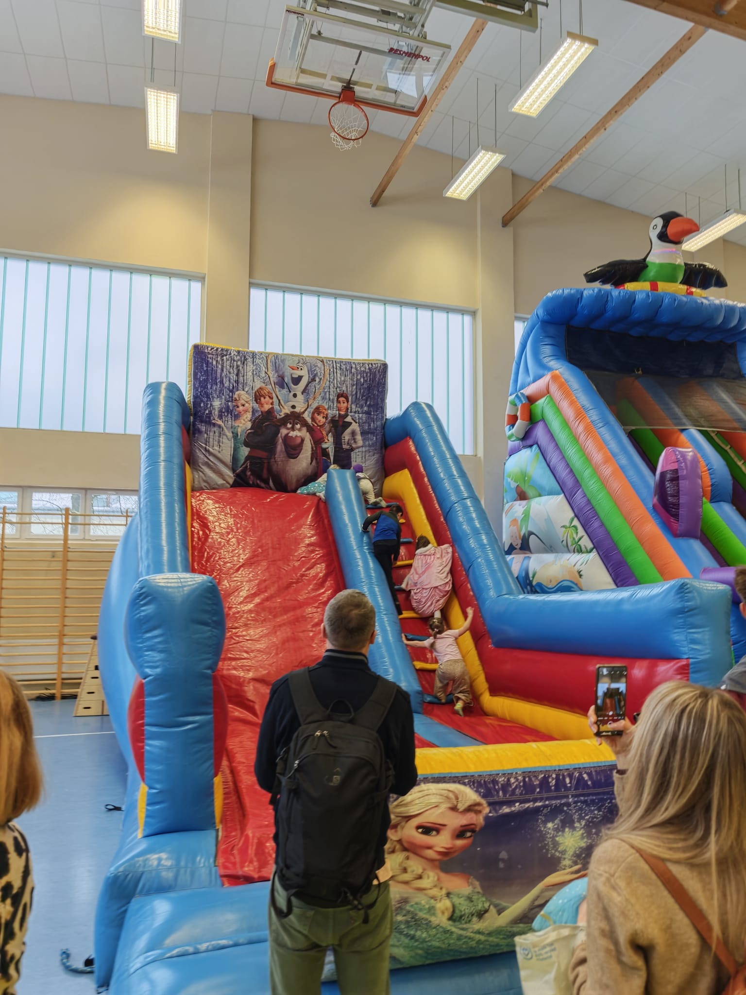 Kolorowy dmuchany zamek z motywem 'Kraina Lodu' w hali sportowej. Dzieci wspinają się po schodach, a dorośli robią zdjęcia. Na szczycie zjeżdżalni duży, dmuchany tukan.