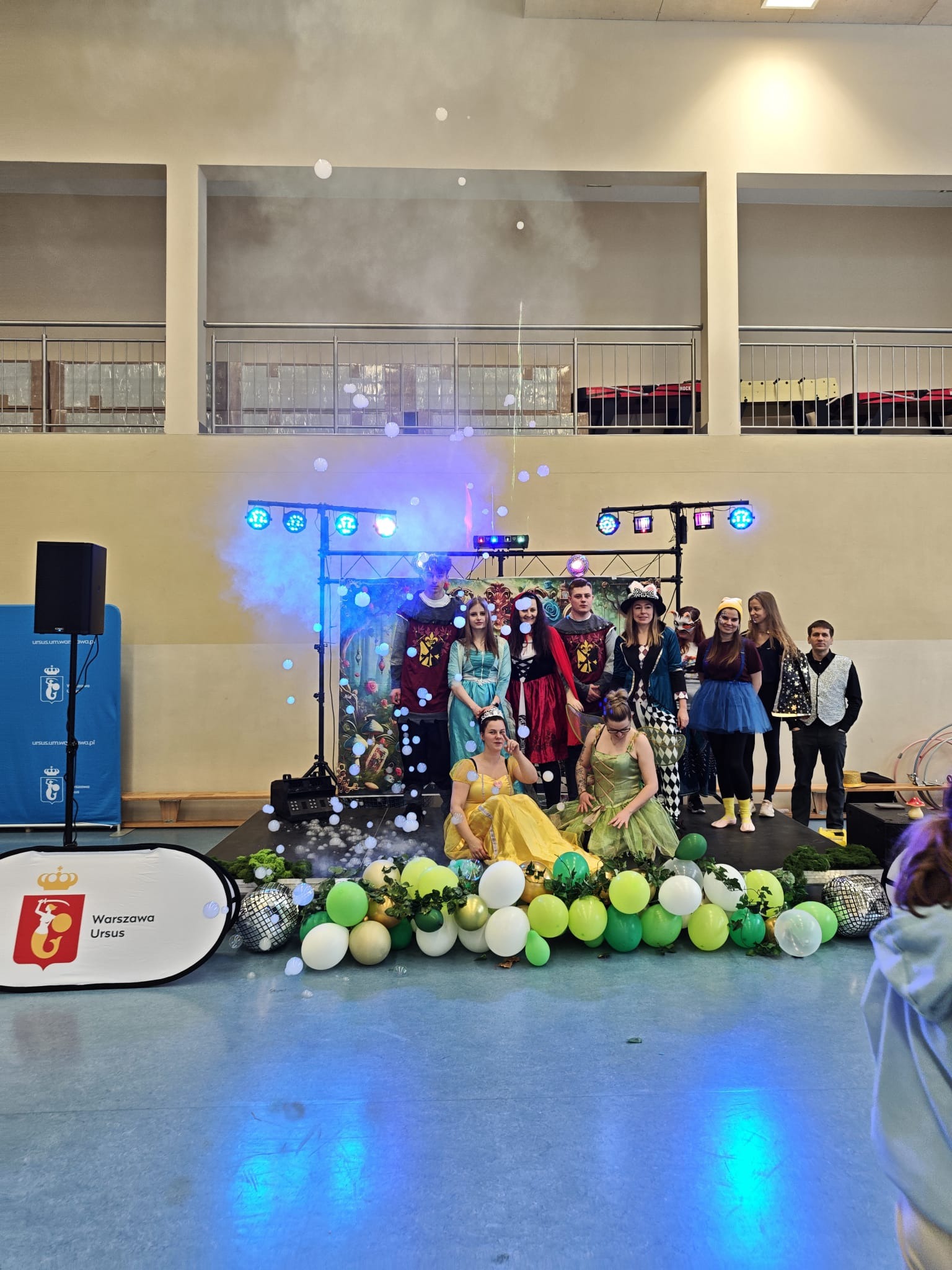 Scena z grupą osób w kostiumach, oświetlona reflektorami i otoczona dekoracjami z balonów i banerem Warszawa Ursus. Efekty specjalne w postaci baniek mydlanych.