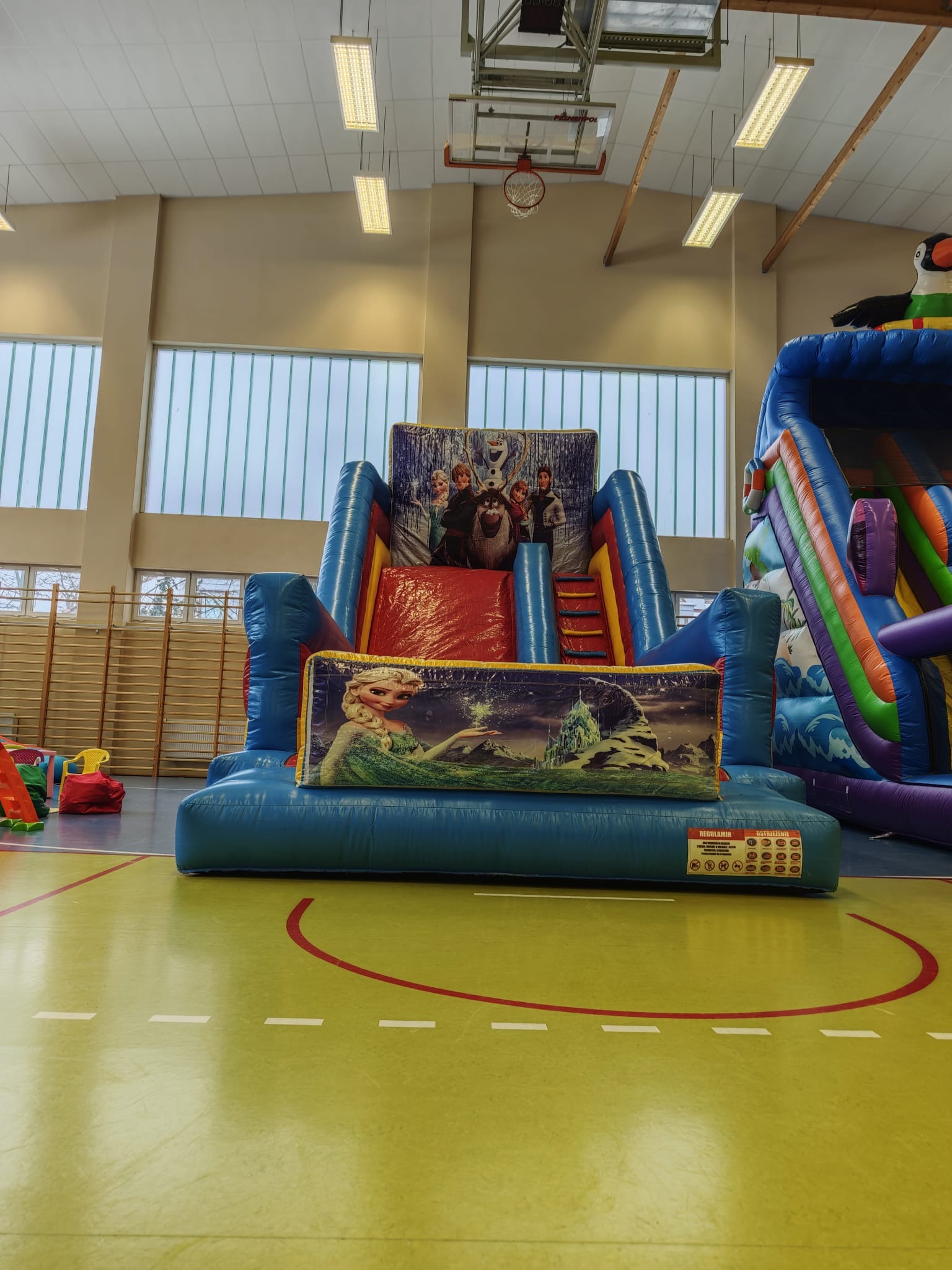Duży, niebieski dmuchany zamek z motywem 'Kraina Lodu' w hali sportowej. Widoczne okna i parkiet. Na drugim planie kolorowa dmuchana zjeżdżalnia z motywem tukana.