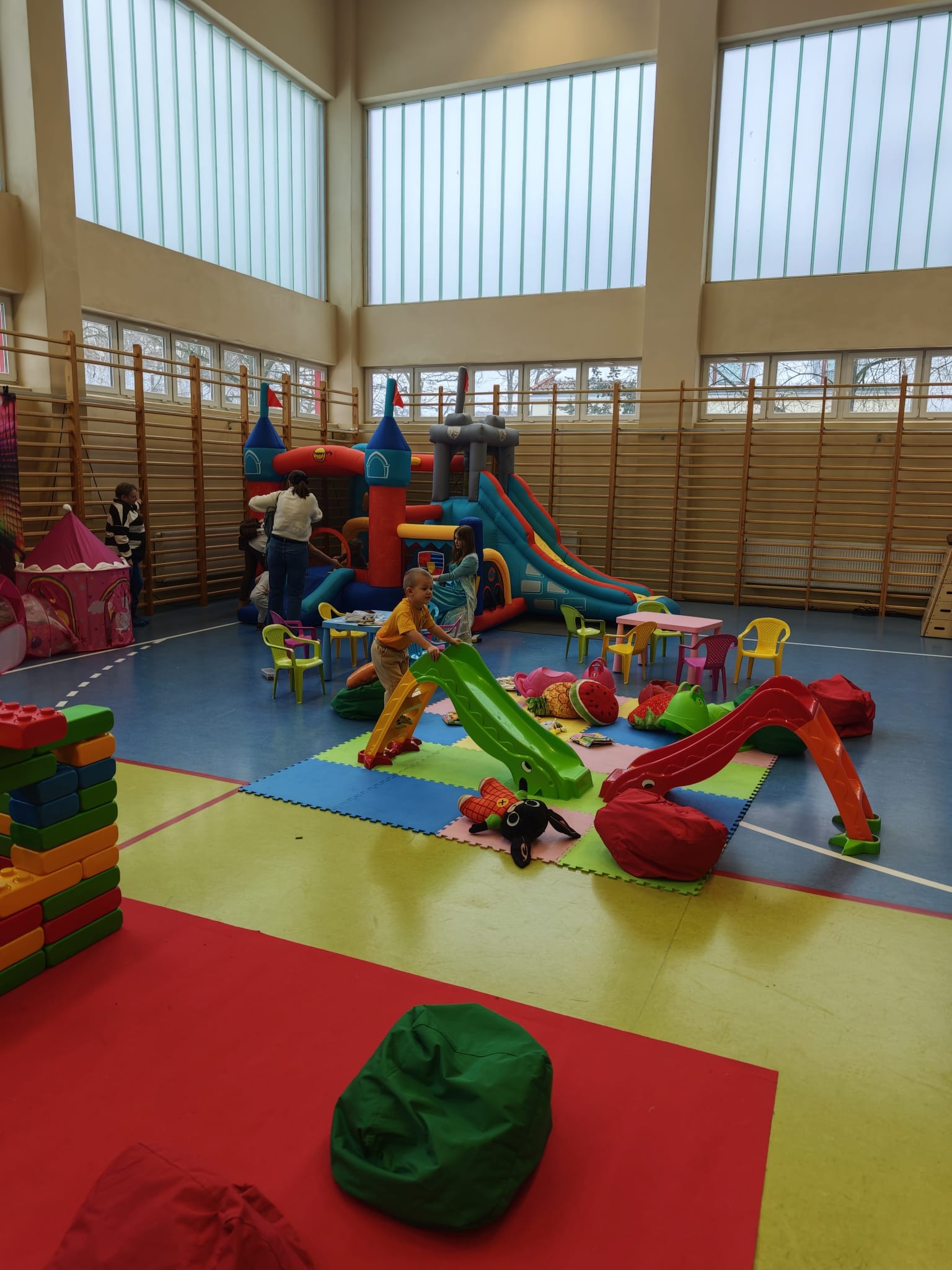 Sala zabaw w hali sportowej z dmuchanym zamkiem, zjeżdżalniami, stolikami i pufami. Dzieci bawią się na kolorowej podłodze z puzzli.