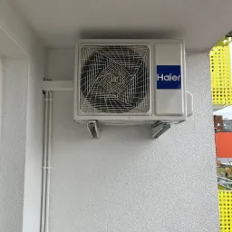 Biała jednostka zewnętrzna klimatyzacji marki Haier zamontowana na ścianie budynku z widocznymi przewodami w białych listwach i fragmentem żółtej balustrady balkonu w tle.