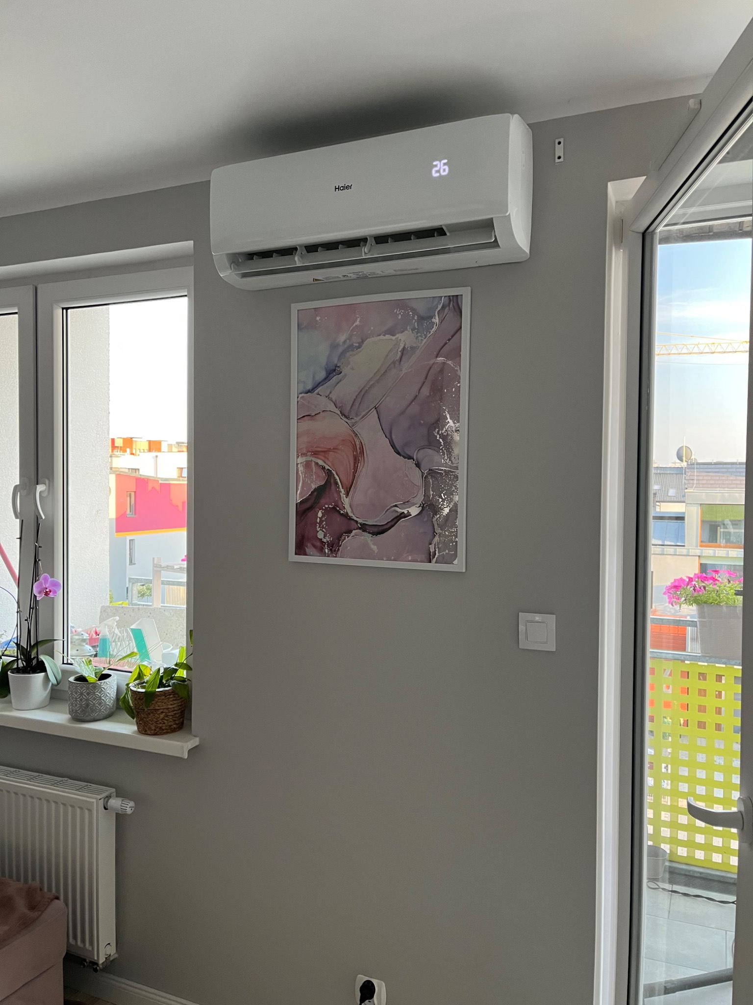 Biała jednostka klimatyzacji Haier zamontowana na szarej ścianie nad obrazem w ramce, z widocznym wyświetlaczem temperatury 26 stopni Celsjusza, obok okna z kwiatami na parapecie i wyjściem...