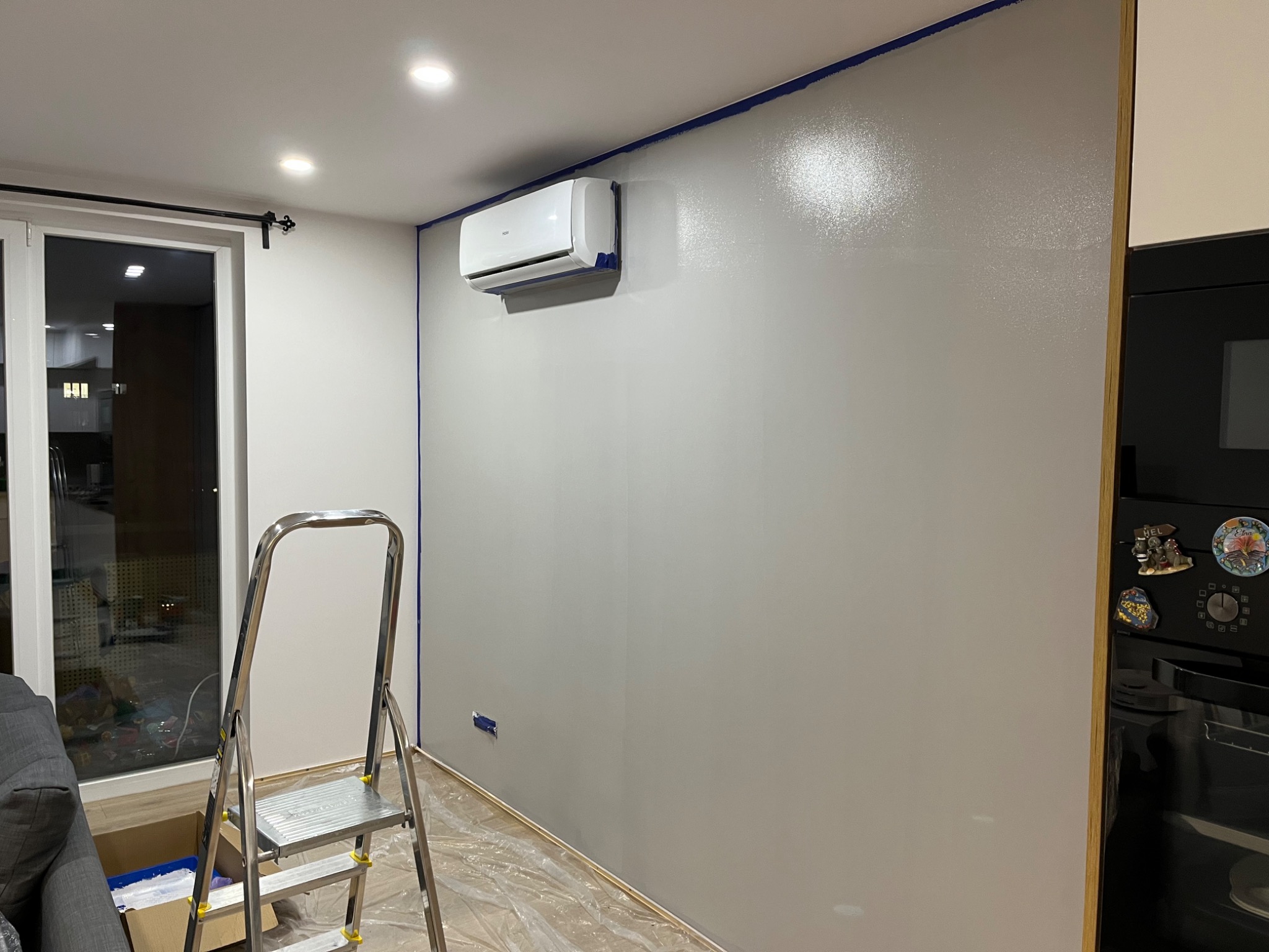 Świeżo pomalowana szara ściana z zamontowanym klimatyzatorem Haier, zabezpieczona niebieską taśmą malarską, z rozłożoną folią ochronną na podłodze i metalową drabiną w pomieszczeniu.