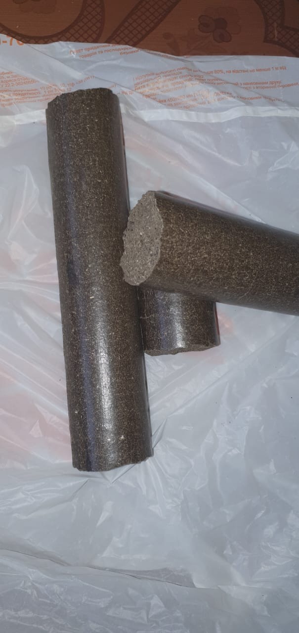 Trzy cylindryczne kawałki brązowego paliwa stałego na białym, pomarszczonym tle. Jeden kawałek stoi pionowo, a dwa leżą obok siebie, z widoczną fakturą cięcia jednego z nich.