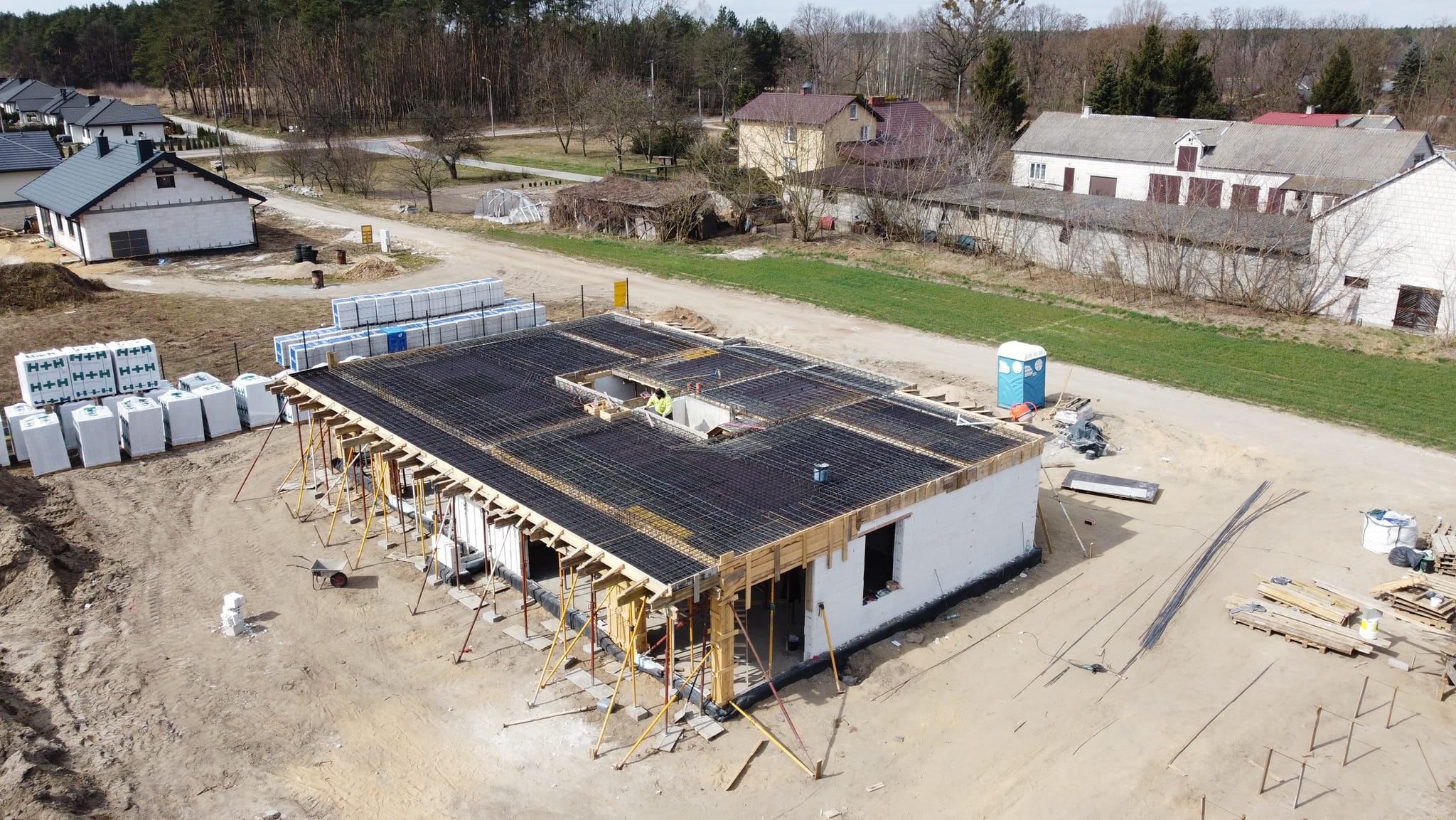 Budowa domu murowanego w Serocku: widok z góry na strop w trakcie zbrojenia, widoczne szalunki, stalowe pręty zbrojeniowe, pracownik w żółtej kamizelce w otworze stropowym oraz palety z bloczkami H+H.