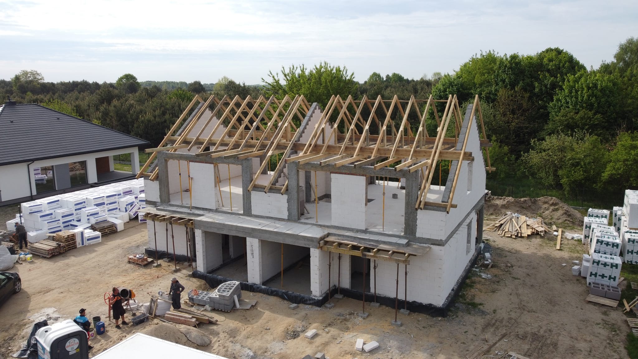 Wznoszenie dwukondygnacyjnego domu murowanego z białych bloczków, widoczna drewniana więźba dachowa, materiały budowlane składowane obok, pracownicy w trakcie wykonywania prac murarskich.