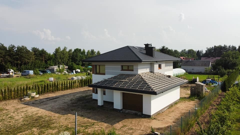 Dwukondygnacyjny dom z szarą elewacją i czarnym dachem w trakcie budowy, widok z góry na częściowo ukończony dach nad garażem, otoczony zielenią i drzewami.