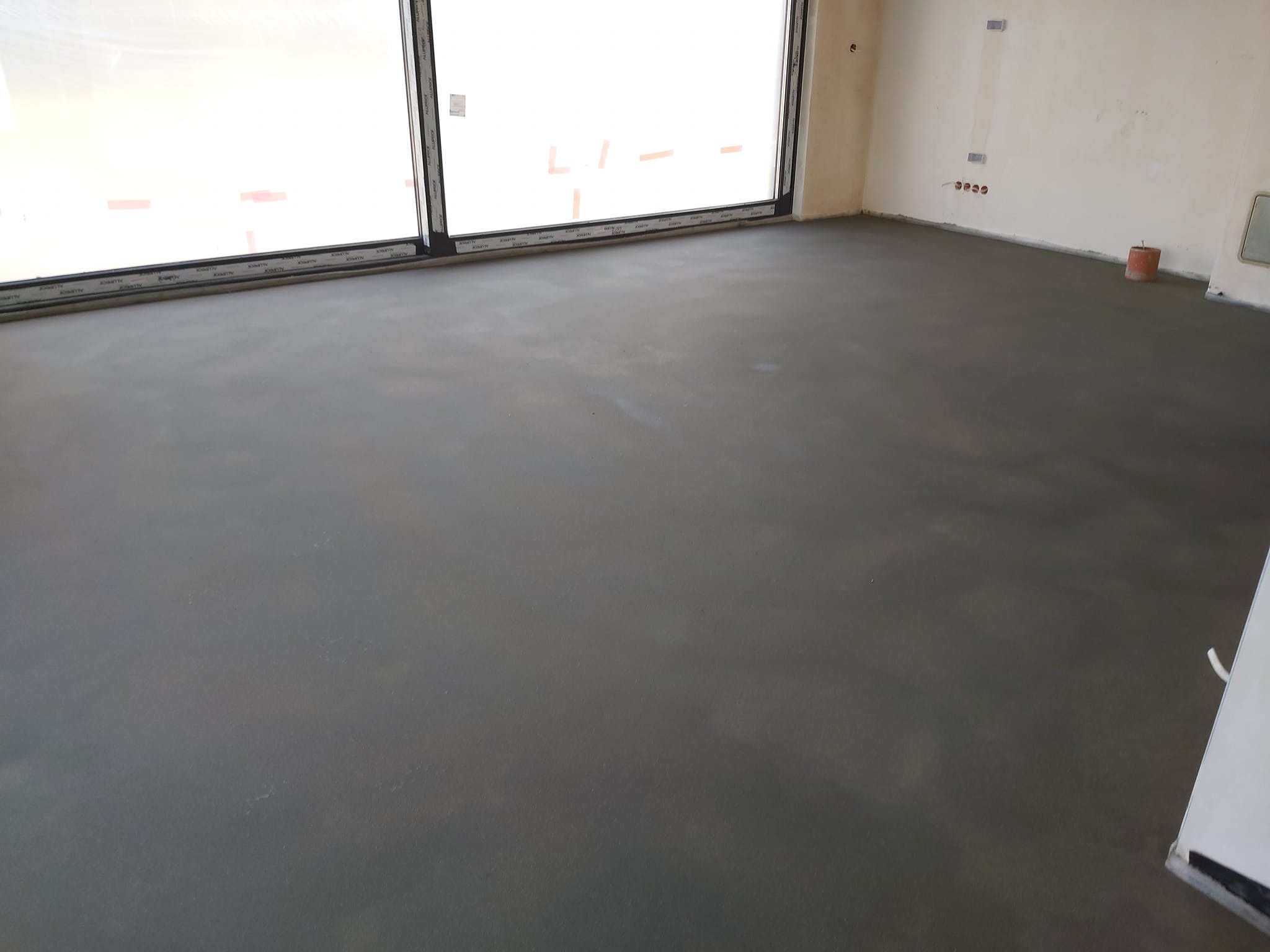 Świeżo wylany, gładki jastrych cementowy w jasnym pomieszczeniu z dużym oknem balkonowym. Widoczne ślady po narzędziach i wiaderko po prawej stronie.