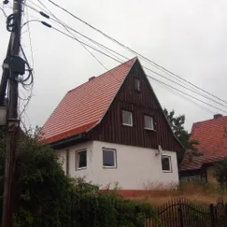 Dwupiętrowy dom z czerwonym dachem dwuspadowym, białymi ścianami na parterze i brązowym drewnianym wykończeniem na piętrze, widoczny zza zarośli i żelaznego ogrodzenia w pochmurny dzień.