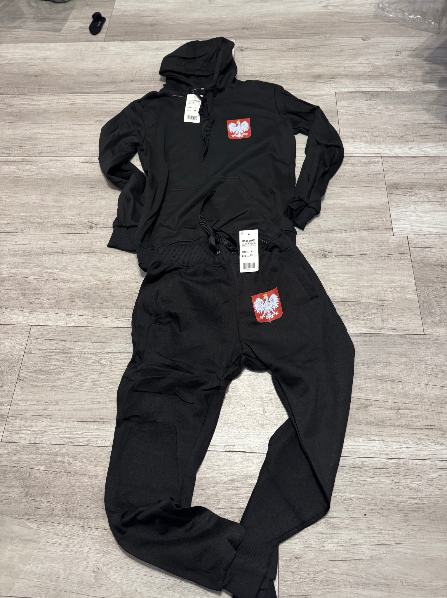 Czarny dres (bluza z kapturem i spodnie) z naszytym godłem Polski leży na panelach podłogowych. Widoczne metki.