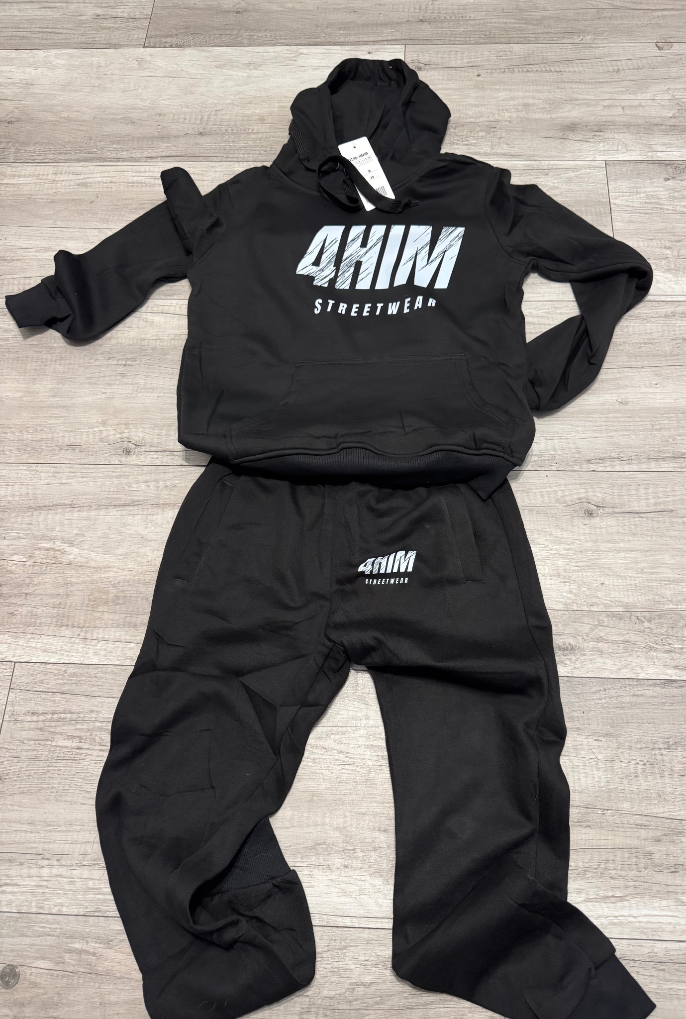 Czarny dres z kapturem, z białym nadrukiem '4HIM STREETWEAR', leżący na podłodze z paneli. Widoczna metka. Całość w stylu streetwear.