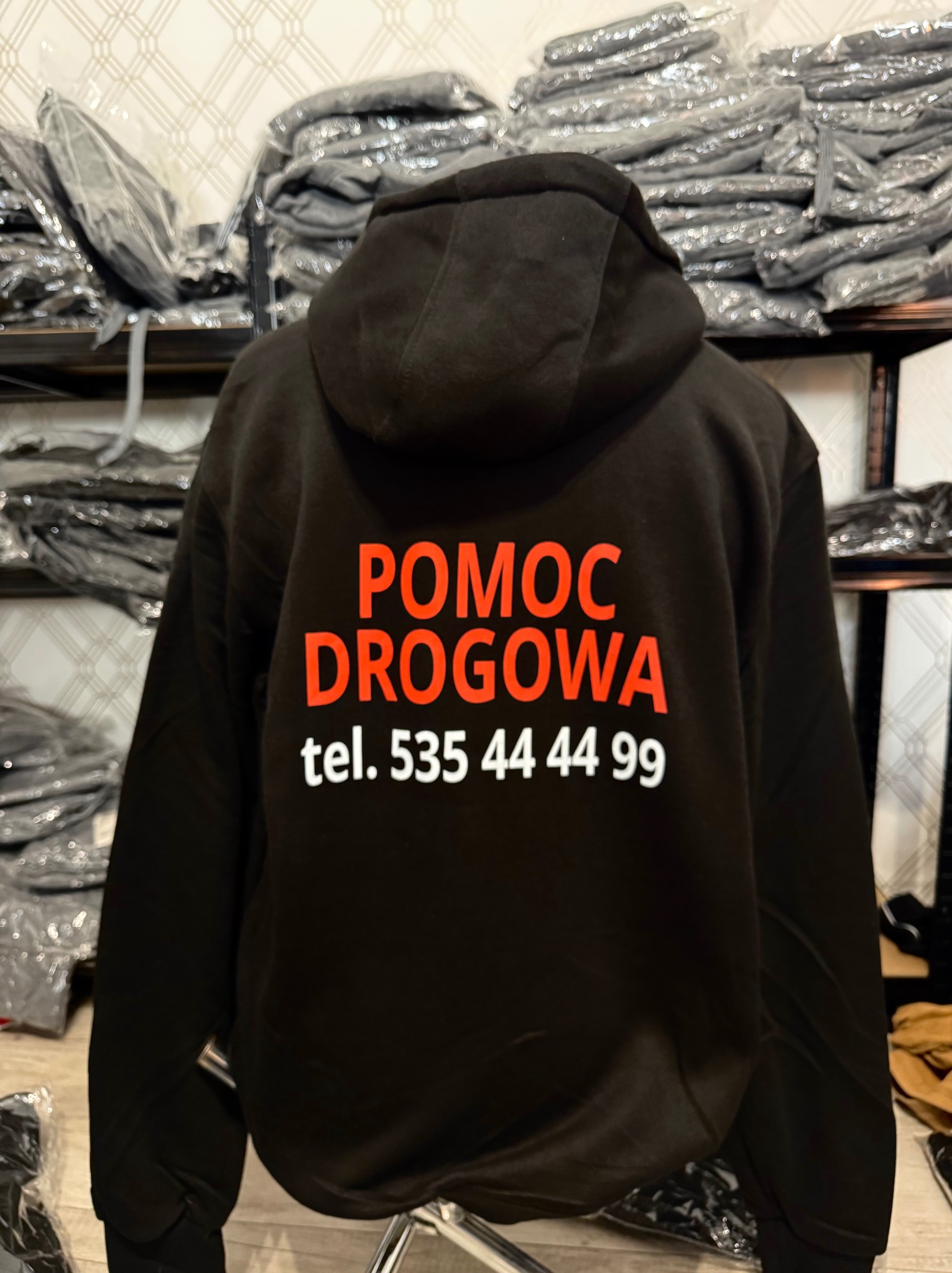 Czarna bluza z kapturem z nadrukiem 'Pomoc Drogowa tel. 535 44 44 99' w kolorze pomarańczowym, prezentowana na manekinie, w tle stosy zapakowanych bluz.