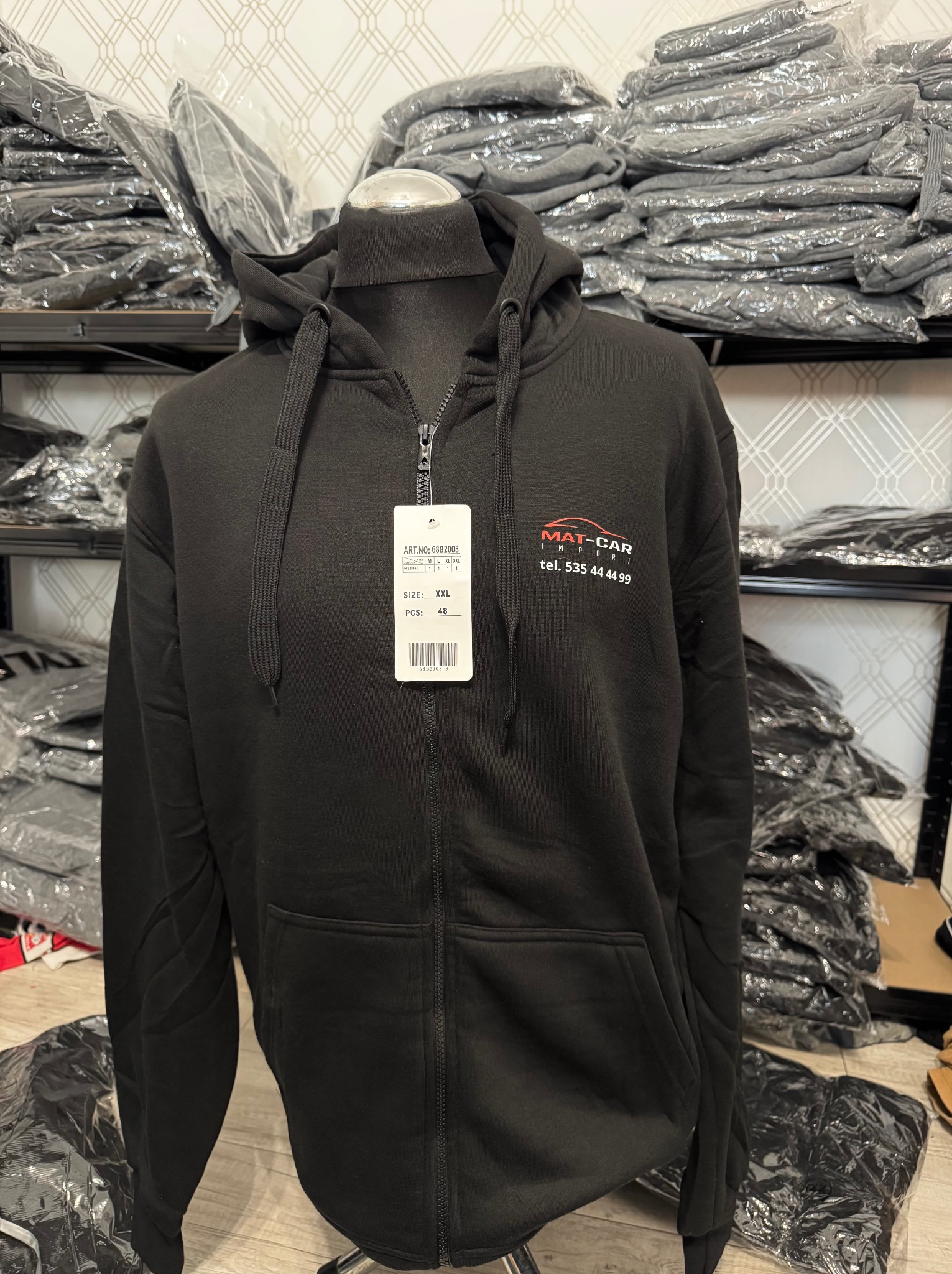 Czarna bluza z kapturem na manekinie, z logo 'MAT-CAR' i numerem telefonu. W tle stosy bluz w foliowych opakowaniach na metalowych półkach.