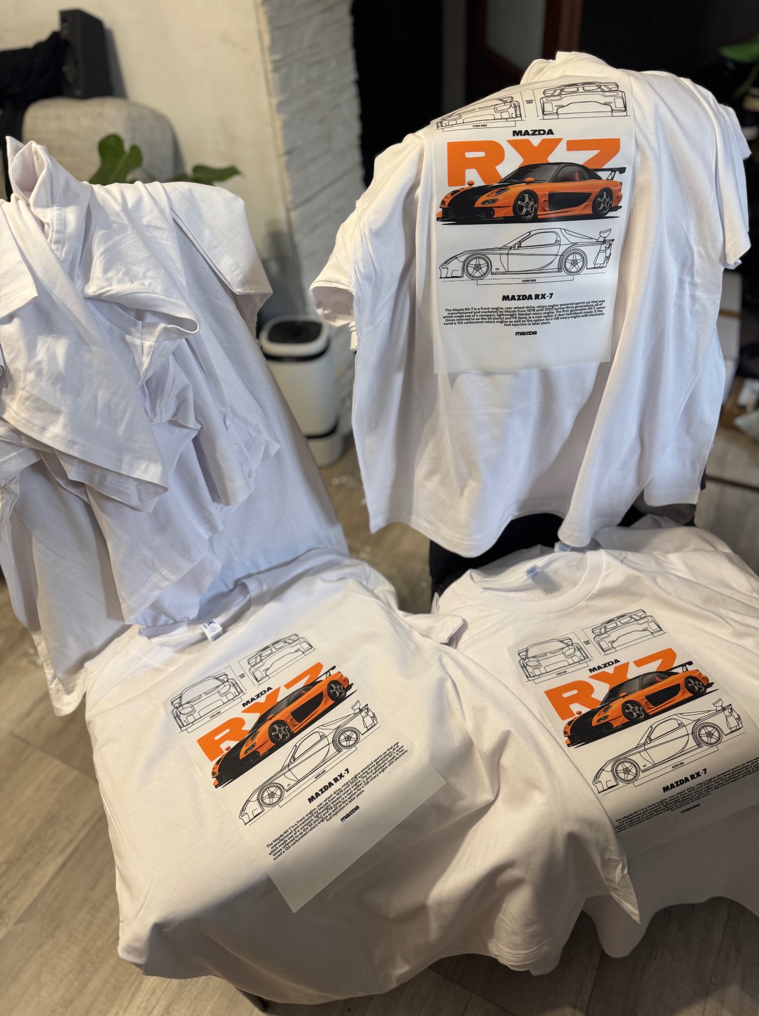 Białe koszulki z nadrukiem pomarańczowo-czarnego auta Mazda RX-7 z technicznym schematem. Kilka sztuk leży na sobie, tworząc stos.