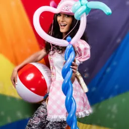 Uśmiechnięta kobieta w różowo-białej sukience w kratę i kapeluszu, trzymająca balon w kształcie serca i długi skręcony niebieski balon, z piłką plażową w dłoni, na tle tęczowej tkaniny.