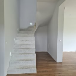 FHU Agan - Surowe betonowe schody z wbudowanym oświetleniem LED na białej ścianie, brązowa podłoga imitująca drewno, widok na jasne wnętrze domu.