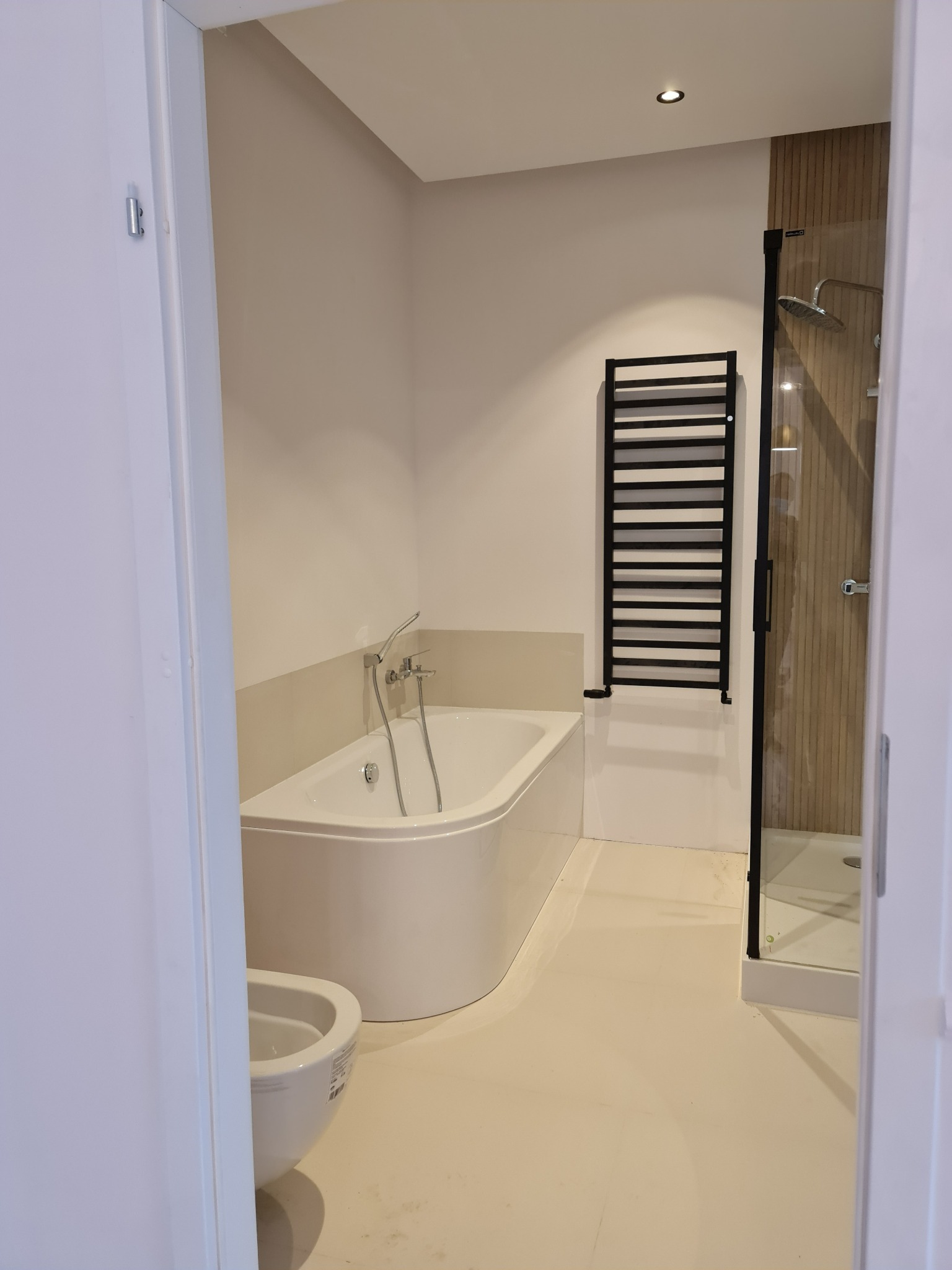 Nowoczesna łazienka z białą wanną, bidetem i kabiną prysznicową z czarną ramą, uzupełniona czarnym grzejnikiem drabinkowym, widok zza otwartych drzwi.