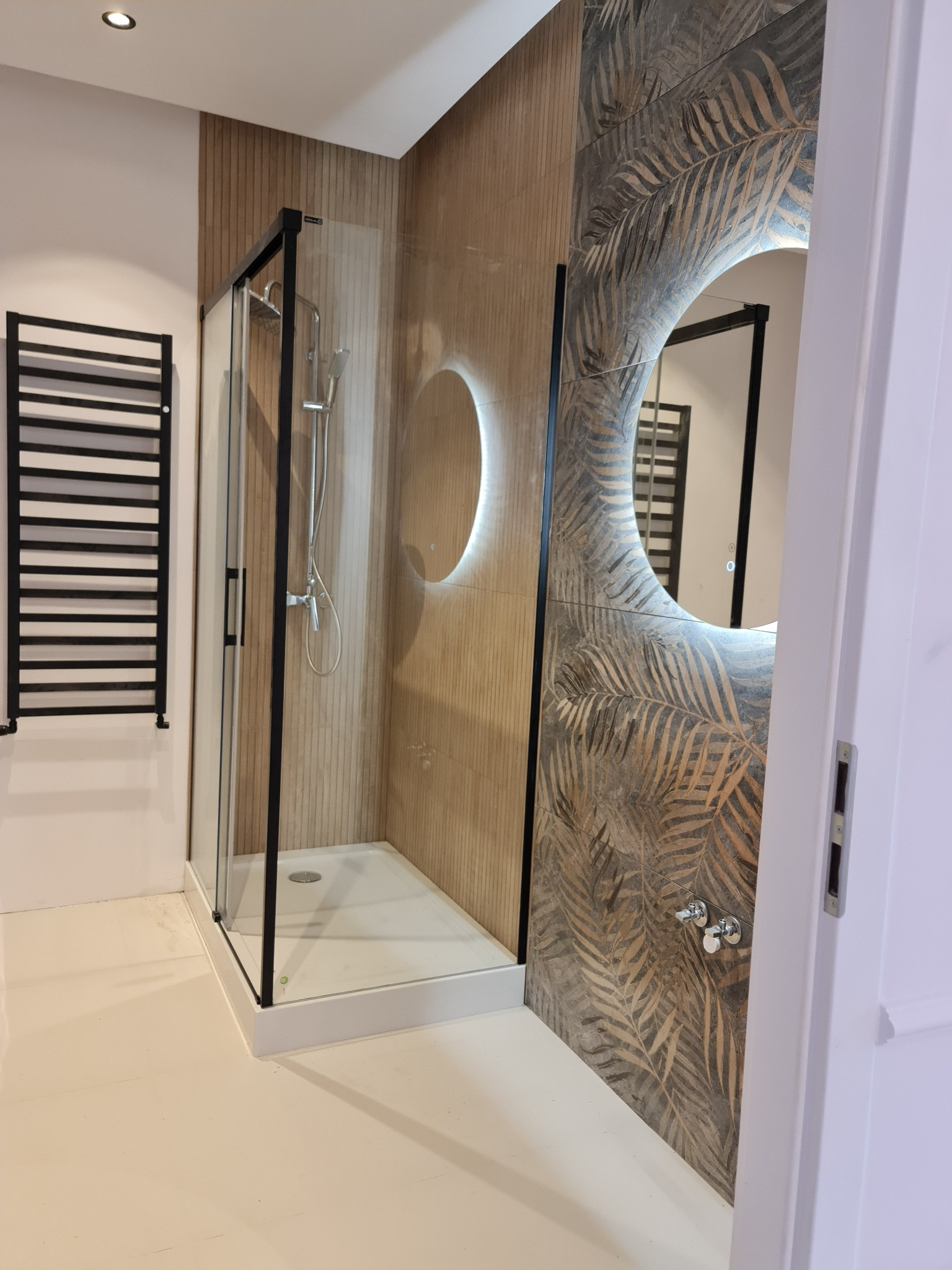 Nowoczesna łazienka z kabiną prysznicową w czarnej ramie, grzejnikiem drabinkowym, okrągłym lustrem z podświetleniem i płytkami z motywem liści palmowych.